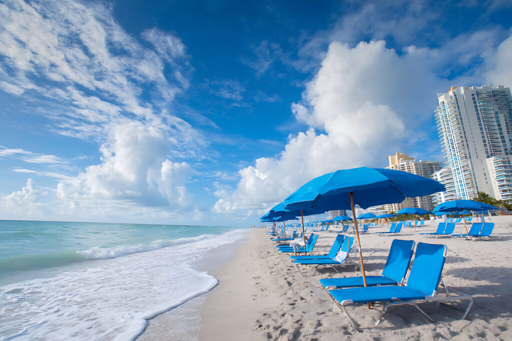 Blue umbrellas and chairs on Miami Beach with ocean waves and beachfront buildings.