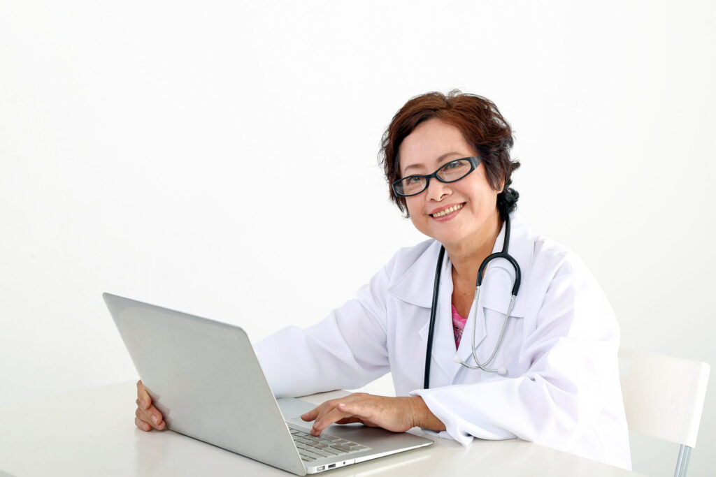 Female doctor wearing a white lab coat and stethoscope, smiling while using a laptop at a desk in a medical office.